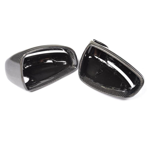 Two black, glossy car mirror covers are lying side by side on a white surface, showcasing their smooth, reflective carbon fiber texture.