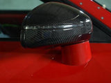 A carbon fiber car side mirror, mounted on a bright red vehicle, reflects light with a sleek, aerodynamic shape in an indoor setting.
