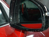 A car's side mirror with a carbon fiber frame reflects an obscured view, attached to a glossy red vehicle body in a garage or showroom setting.
