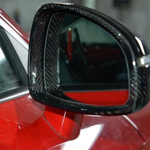 A car's side mirror with a carbon fiber frame reflects an obscured view, attached to a glossy red vehicle body in a garage or showroom setting.