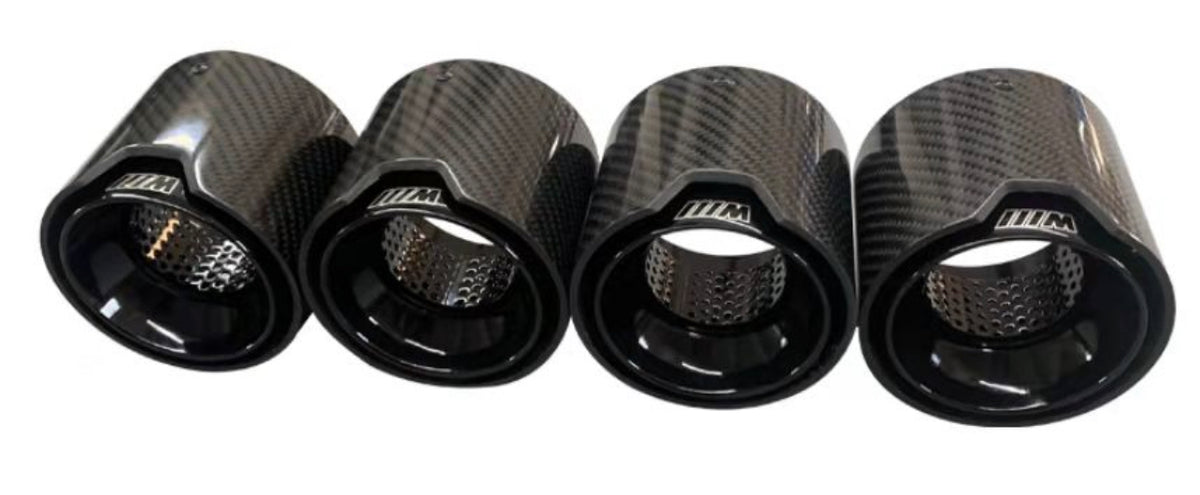 Four carbon-fiber exhaust tips display a glossy finish and perforated interiors. They are aligned horizontally on a plain, white background. Each tip has "///M" inscribed on it.