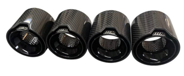 Four carbon-fiber exhaust tips display a glossy finish and perforated interiors. They are aligned horizontally on a plain, white background. Each tip has "///M" inscribed on it.