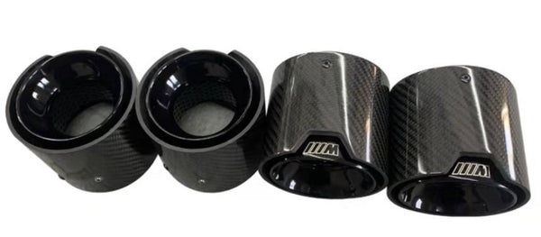 Four carbon fiber exhaust tips, two with "///M" logo, resting on a smooth surface. They feature a glossy finish and a textured design, emphasizing a sleek automotive accessory context.