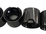 Four glossy carbon-fiber exhaust tips, aligned side by side, featuring a textured finish and each engraved with a stylized “///M” logo on the rim, against a plain background.