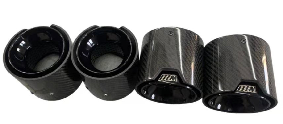Four glossy carbon-fiber exhaust tips, aligned side by side, featuring a textured finish and each engraved with a stylized “///M” logo on the rim, against a plain background.