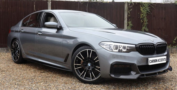 A gray sedan is parked on gravel, featuring sleek black wheels and illuminated headlights. The license plate reads "CARBON ACCENTS," with a wooden fence in the background.