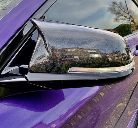 Car side mirror reflects nearby houses, mounted on a vibrant purple vehicle in a residential area. The mirror surface features a glossy, black finish.