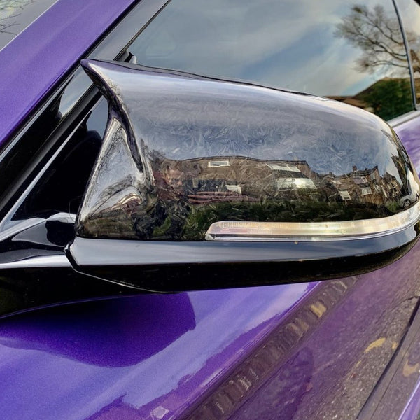A black side mirror of a purple car reflects nearby houses and trees on a clear day, emphasizing the surrounding residential area.