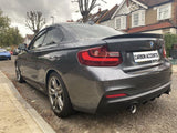 A gray BMW coupe is parked on a residential street. Bare trees and brick houses line the background. The license plate reads 