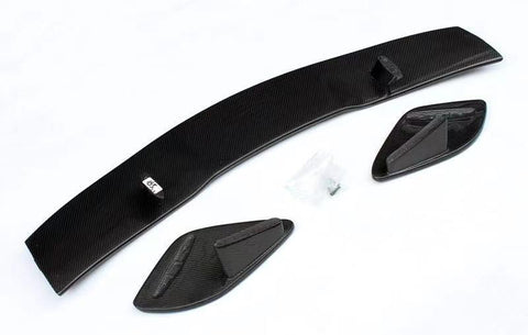 Carbon fiber car spoiler components lie on a white surface, consisting of a long curved piece and two smaller side elements. A small bag containing screws is nearby.