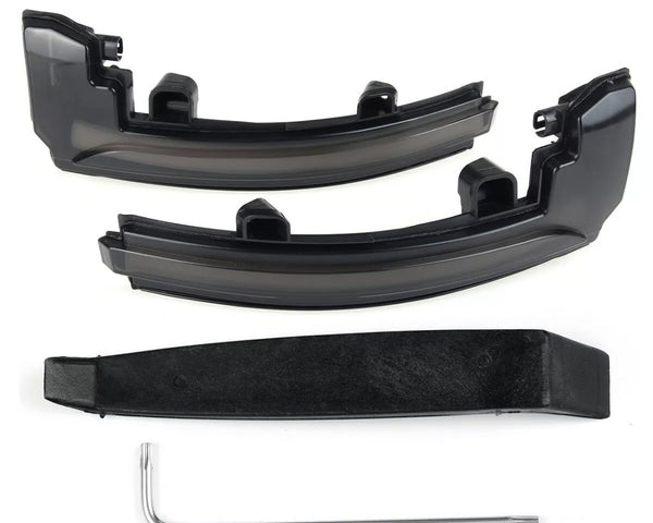 Two black plastic car light cover pieces rest on a white surface, accompanied by a black mounting bracket and a silver L-shaped tool beneath them.