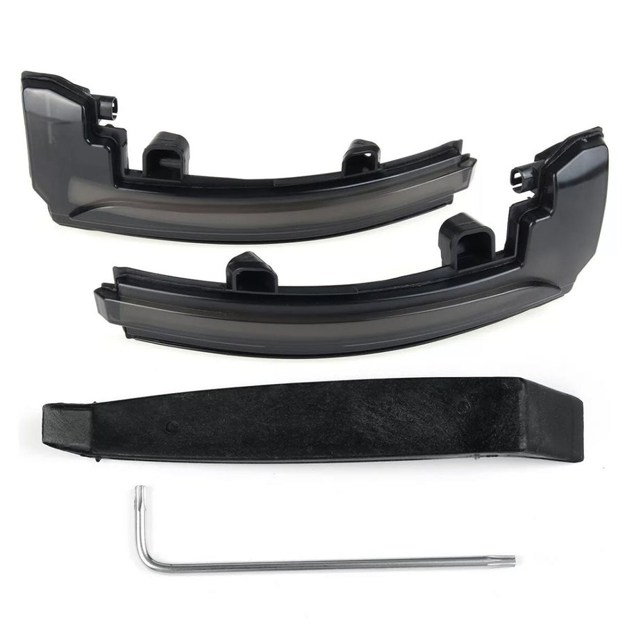 Two black plastic car light cover pieces rest on a white surface, accompanied by a black mounting bracket and a silver L-shaped tool beneath them.