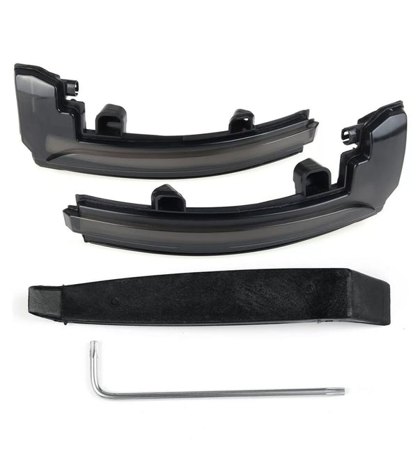 Two black plastic car light cover pieces rest on a white surface, accompanied by a black mounting bracket and a silver L-shaped tool beneath them.