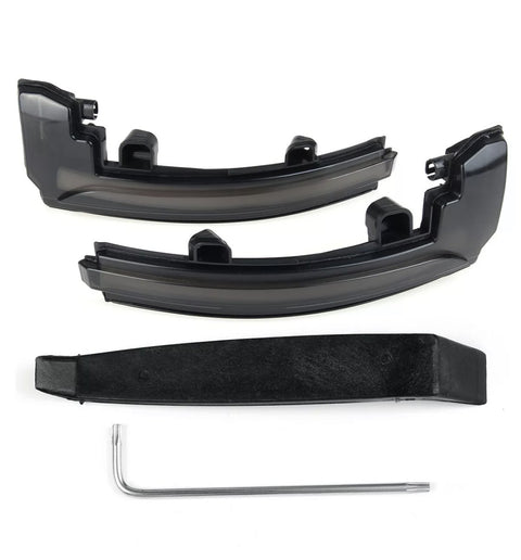 Two black plastic car light cover pieces rest on a white surface, accompanied by a black mounting bracket and a silver L-shaped tool beneath them.