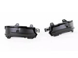 Two black plastic automotive parts, likely fog light housings, are positioned side by side facing outward. They feature mounting brackets and an angular design, set against a plain white background.