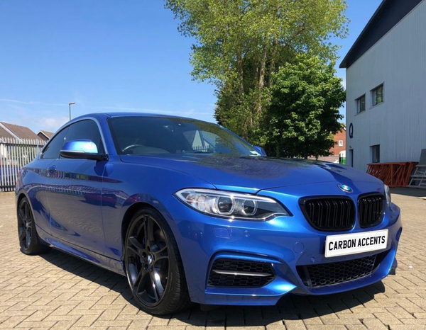 A blue car is parked in a sunny lot, surrounded by trees and buildings. The front license plate reads "CARBON ACCENTS."