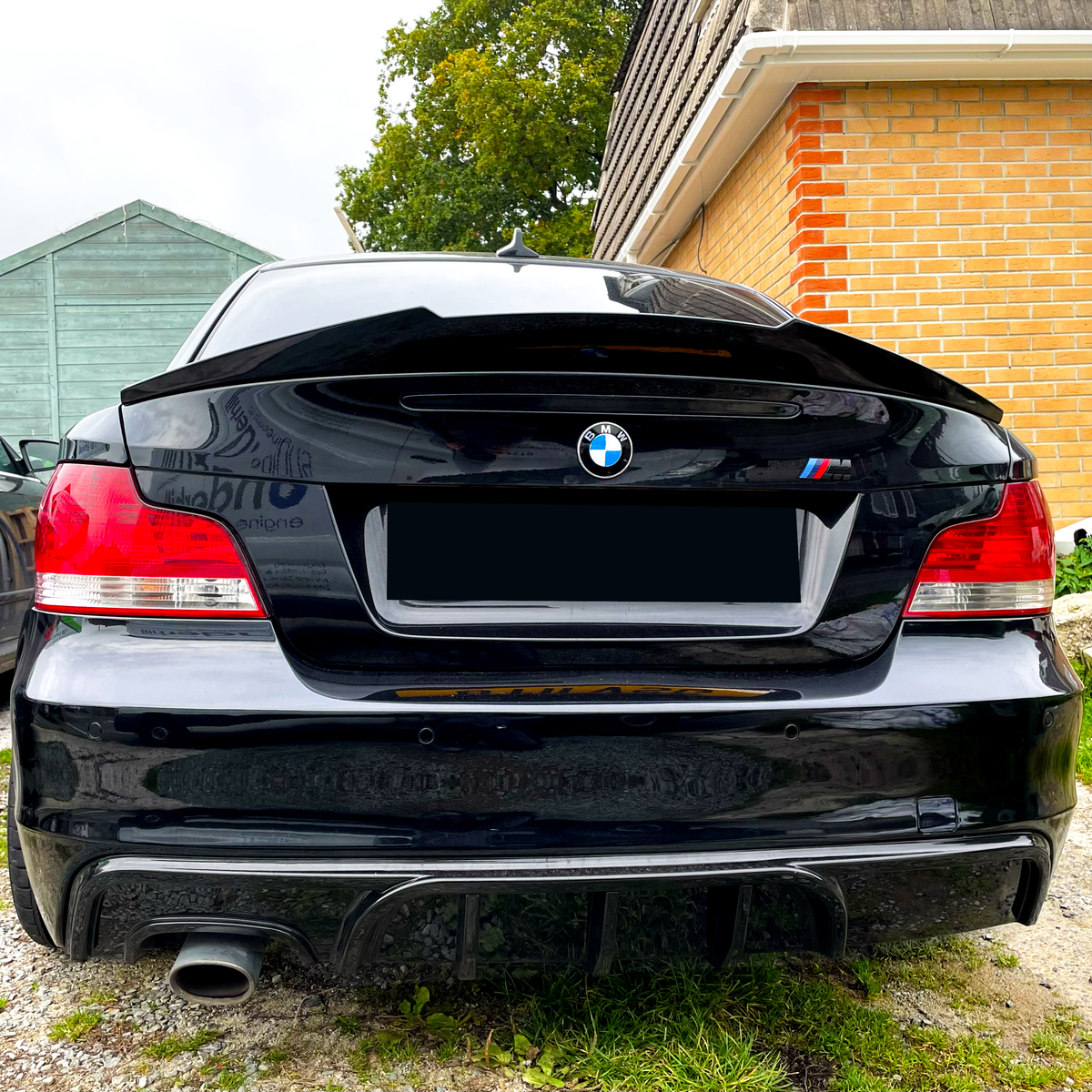 A black BMW car is parked on a gravel driveway beside a brick house and a green wooden shed, with its rear view showing distinct red taillights.