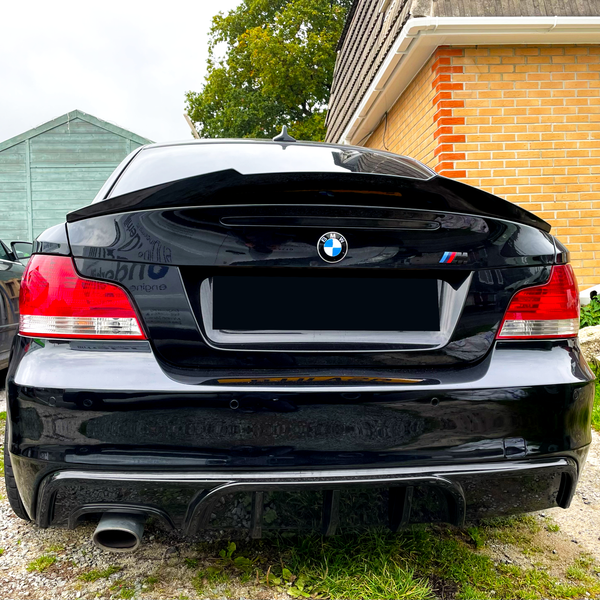 A black BMW car is parked on a gravel driveway beside a brick house and a green wooden shed, with its rear view showing distinct red taillights.