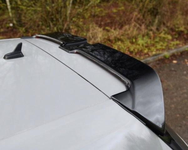 A car roof features a black roof spoiler attached to the rear. The vehicle is parked near a grassy area with trees in the background.