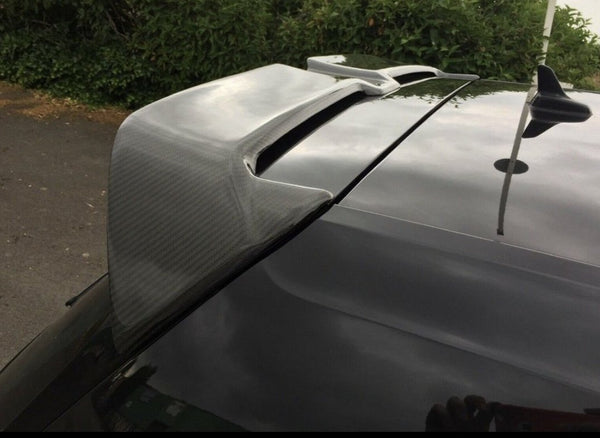 A car's carbon fiber spoiler is parked outdoors, with bushes and a driveway visible in the background.