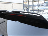 A close-up of a black car's rear spoiler, featuring a red brake light strip, situated outdoors near a light-colored building and a metal fence.