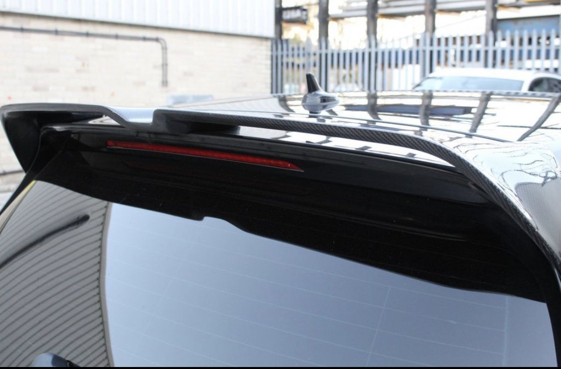 A close-up of a black car's rear spoiler, featuring a red brake light strip, situated outdoors near a light-colored building and a metal fence.