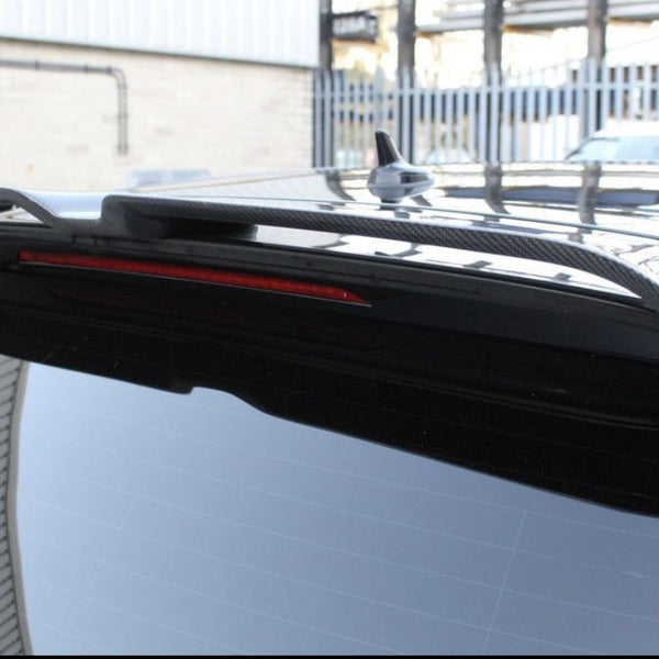 A close-up of a black car's rear spoiler, featuring a red brake light strip, situated outdoors near a light-colored building and a metal fence.