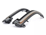 Curved car side mirror indicators, black with translucent housing and amber lights, are positioned on a reflective, white surface.