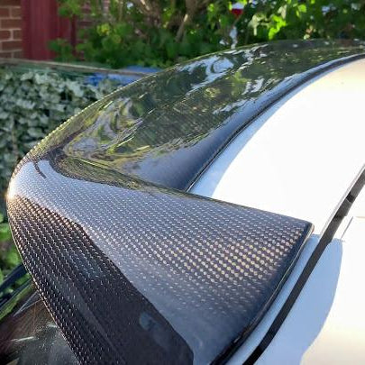 A carbon fiber car spoiler is mounted on a white vehicle, reflecting sunlight. It is situated in a residential driveway with greenery and brickwork visible in the background.