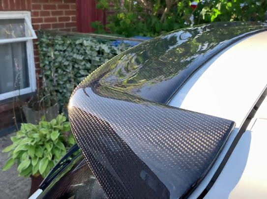 A carbon fiber car spoiler is mounted on a white vehicle, reflecting sunlight. It is situated in a residential driveway with greenery and brickwork visible in the background.