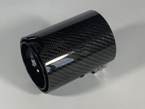 Carbon fiber exhaust tip resting on a white surface, exhibiting a glossy, woven texture. A subtle logo is visible on the edge.
