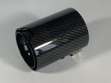 A cylindrical carbon fiber exhaust tip displays reflective surface details, resting on a solid white background. It features a small logo with stylized letters 