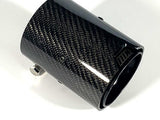 A cylindrical, carbon-fiber car exhaust tip, metallic bolt attached, lies on a white background. A small engraved symbol appears near the upper edge.