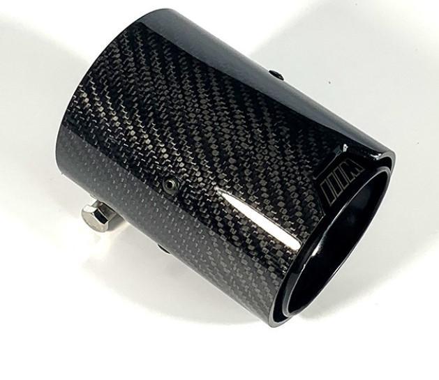 A carbon fiber exhaust tip is lying on a white surface, reflecting light. Its textured, glossy surface and metal clamp indicate it is a vehicle component.