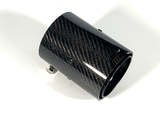 A cylindrical carbon fiber exhaust tip rests on a white surface, featuring a woven pattern and small metal clamp attachments for securing it to a vehicle.