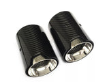 Two cylindrical carbon fiber exhaust tips with shiny metal interiors lie on a white surface. The tips feature a logo resembling 