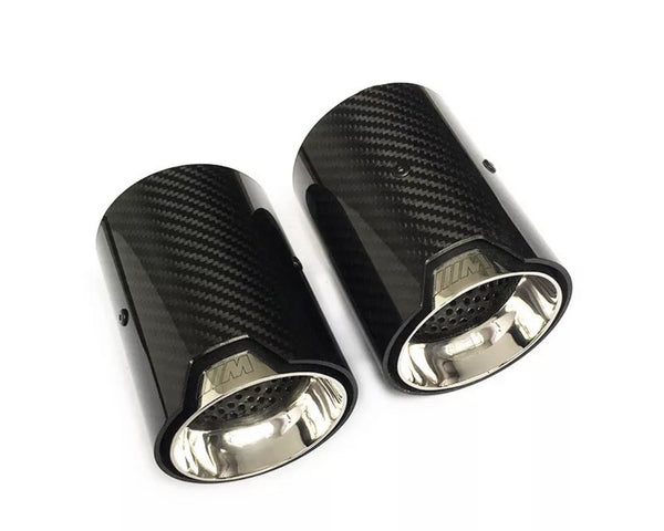 Two cylindrical carbon fiber exhaust tips with shiny metal interiors lie on a white surface. The tips feature a logo resembling "///M" embossed on the outer edge.