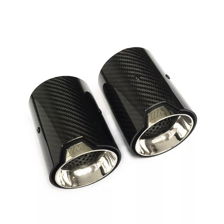Two cylindrical carbon fiber exhaust tips with shiny metal interiors lie on a white surface. The tips feature a logo resembling "///M" embossed on the outer edge.