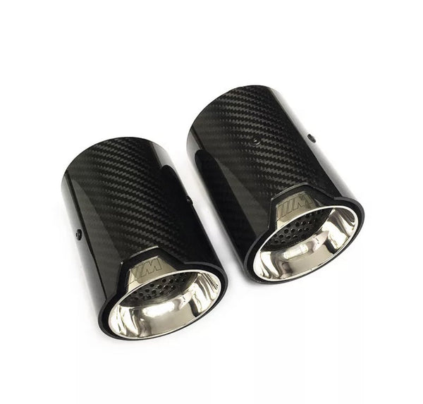Two carbon fiber exhaust tips are displayed side by side, with a glossy black exterior and a metallic interior, on a plain white background. The text "///M" is engraved near the rims.