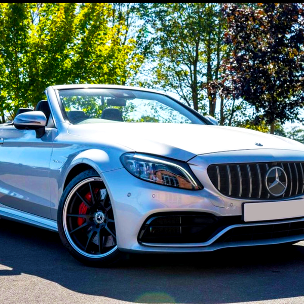 A silver convertible car is parked on pavement, front-facing, under bright sunlight. Trees with green and red leaves create a natural backdrop. The car has visible badges and red brake calipers.