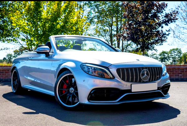 A silver convertible car is parked on pavement, front-facing, under bright sunlight. Trees with green and red leaves create a natural backdrop. The car has visible badges and red brake calipers.