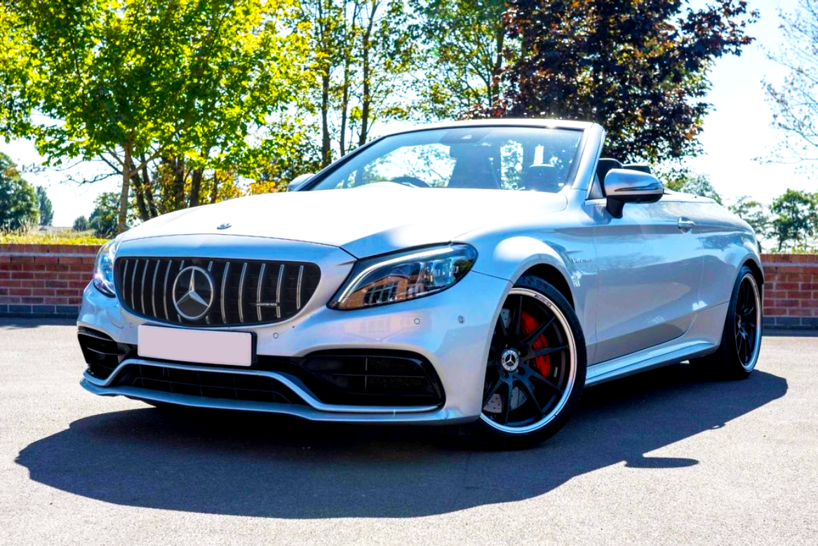 A silver convertible car is parked on a sunny day, showcasing its front grille and sleek design, surrounded by a brick wall and lush green trees.