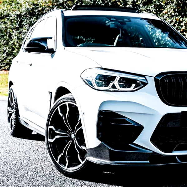 A white SUV is parked on a road with black rims and a sleek design. The background features green foliage under bright sunlight.