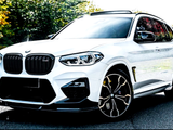 A white BMW X3 M sits parked on a sunlit road, surrounded by lush, green trees. The vehicle features sleek, modern design with prominent front grilles and stylish rims.