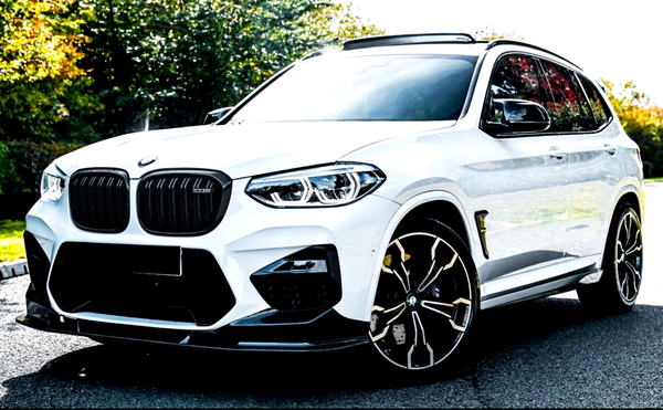 A white BMW SUV is parked on a paved road, displaying a sleek design with distinct alloy wheels and a glossy exterior, surrounded by trees in a sunlit environment.