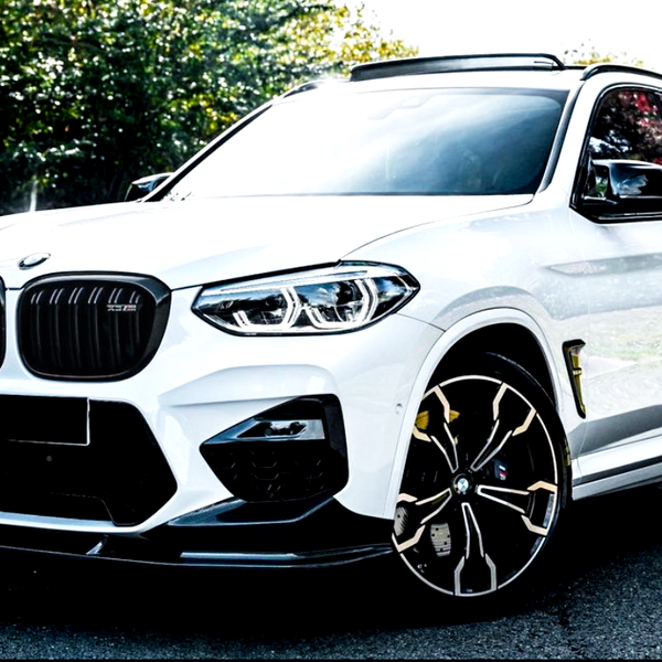 A white BMW SUV parked on a sunlit road, surrounded by green and orange trees. The vehicle features distinctive black grille and large, intricate alloy wheels.