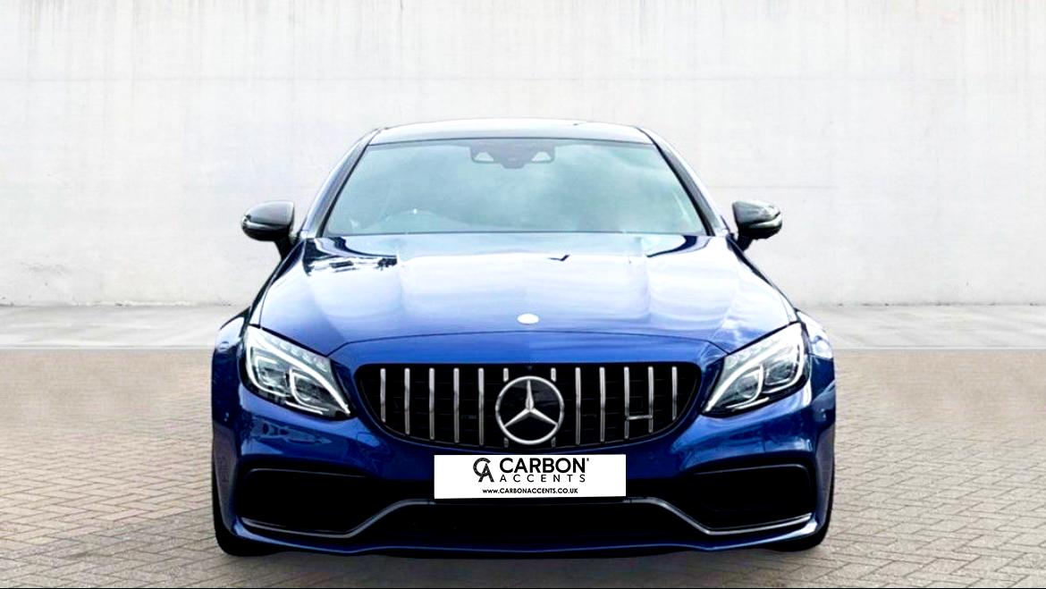 A blue luxury car is parked on a paved surface, facing forward. Its front grille features a prominent emblem. The background is a plain, light-colored wall. Text reads: "CARBON ACCENTS, WWW.CARBONACCENTS.CO.UK."