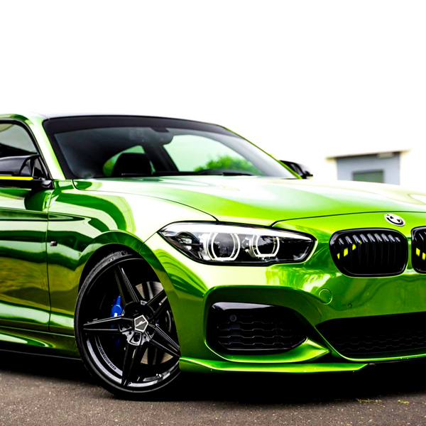 A vibrant green sports car is parked on a paved surface. It features sleek black wheels and distinctive headlights, set against a white building in the background.