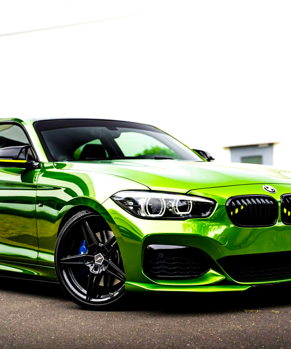 A vibrant green sports car is parked on a paved surface, featuring shiny alloy wheels and a sleek, aerodynamic design, with a blurred, bright backdrop and a visible BMW logo.