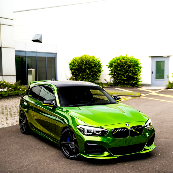 A glossy green car is parked on a paved driveway. Surrounding it are a white building and green shrubs, with a modern entrance visible in the background.